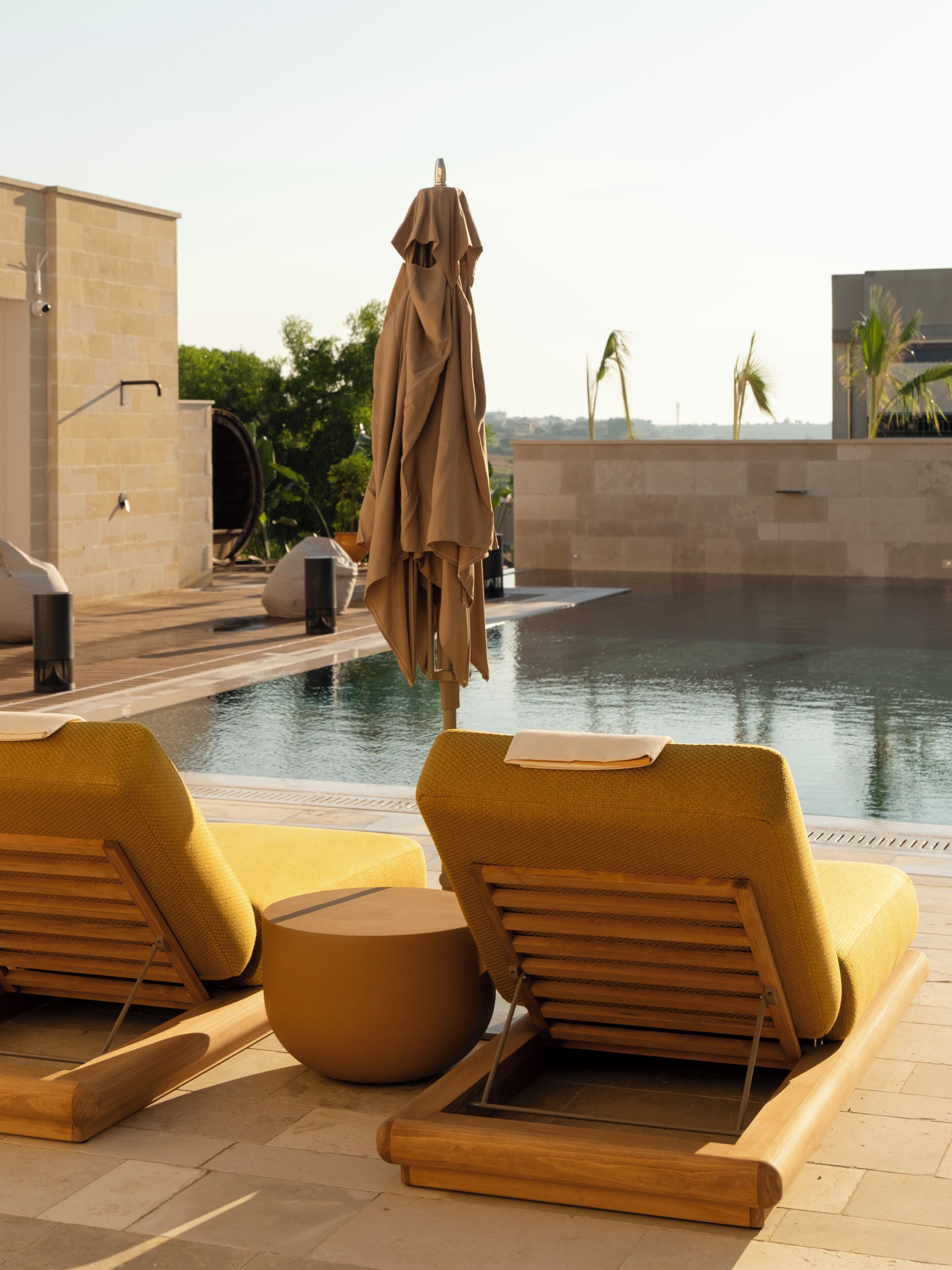 A poolside area with lounge chairs and a covered umbrella, a water feature, and a view of the surrounding landscape.