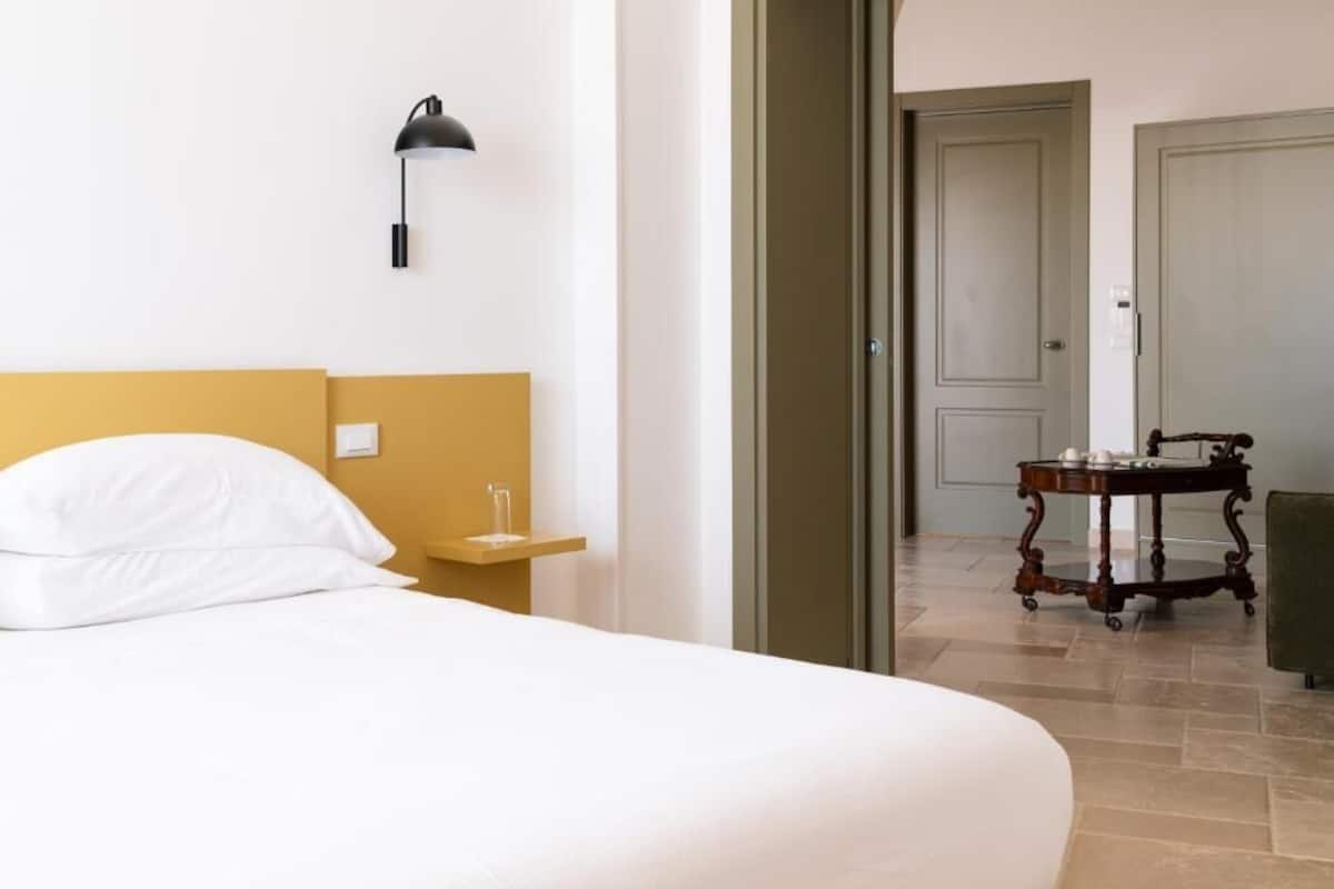 A hotel room with a bed, bedside table, lamp, and a door leading to another room.