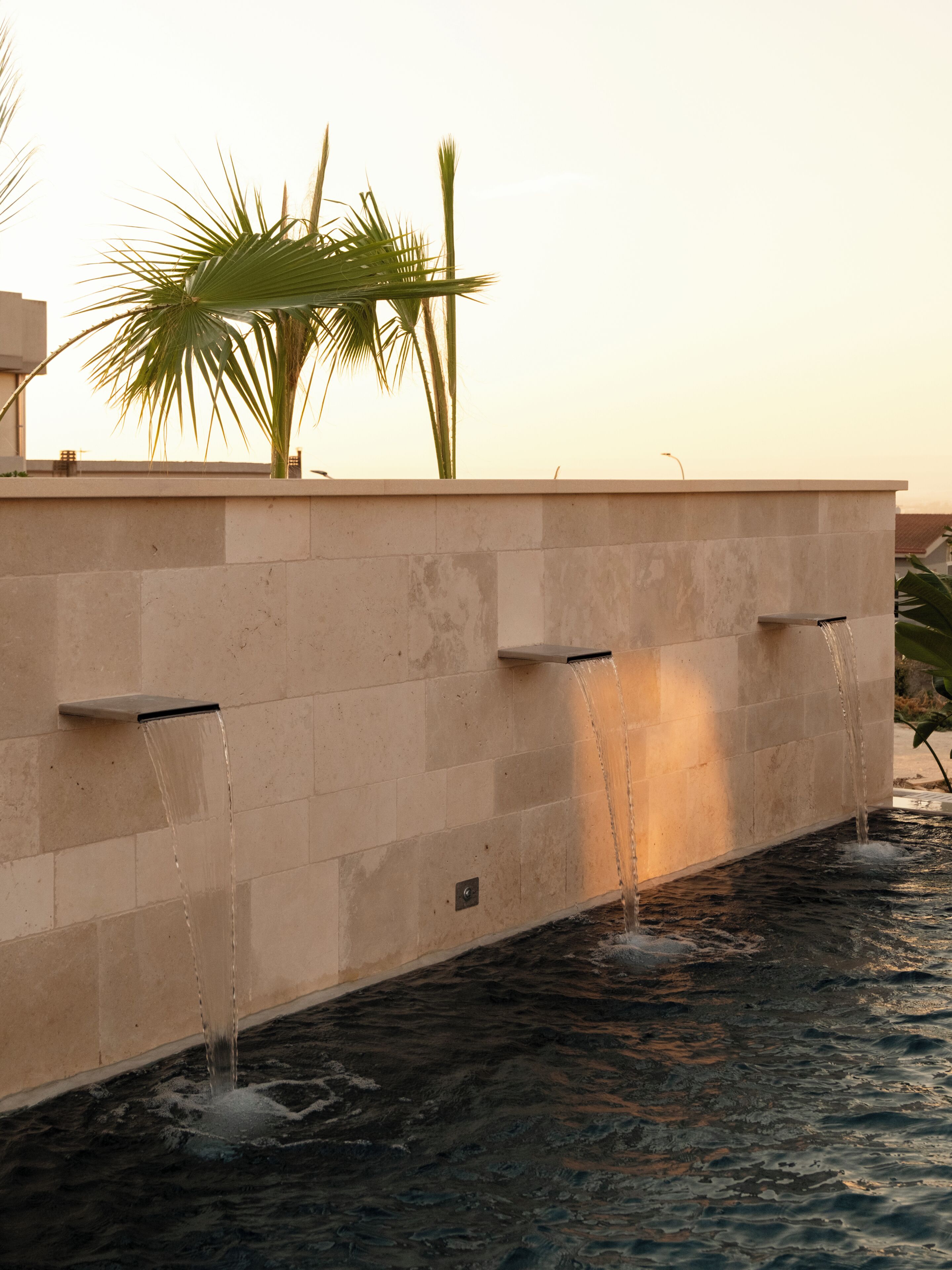 A water feature with multiple streams against a wall, surrounded by palm trees.