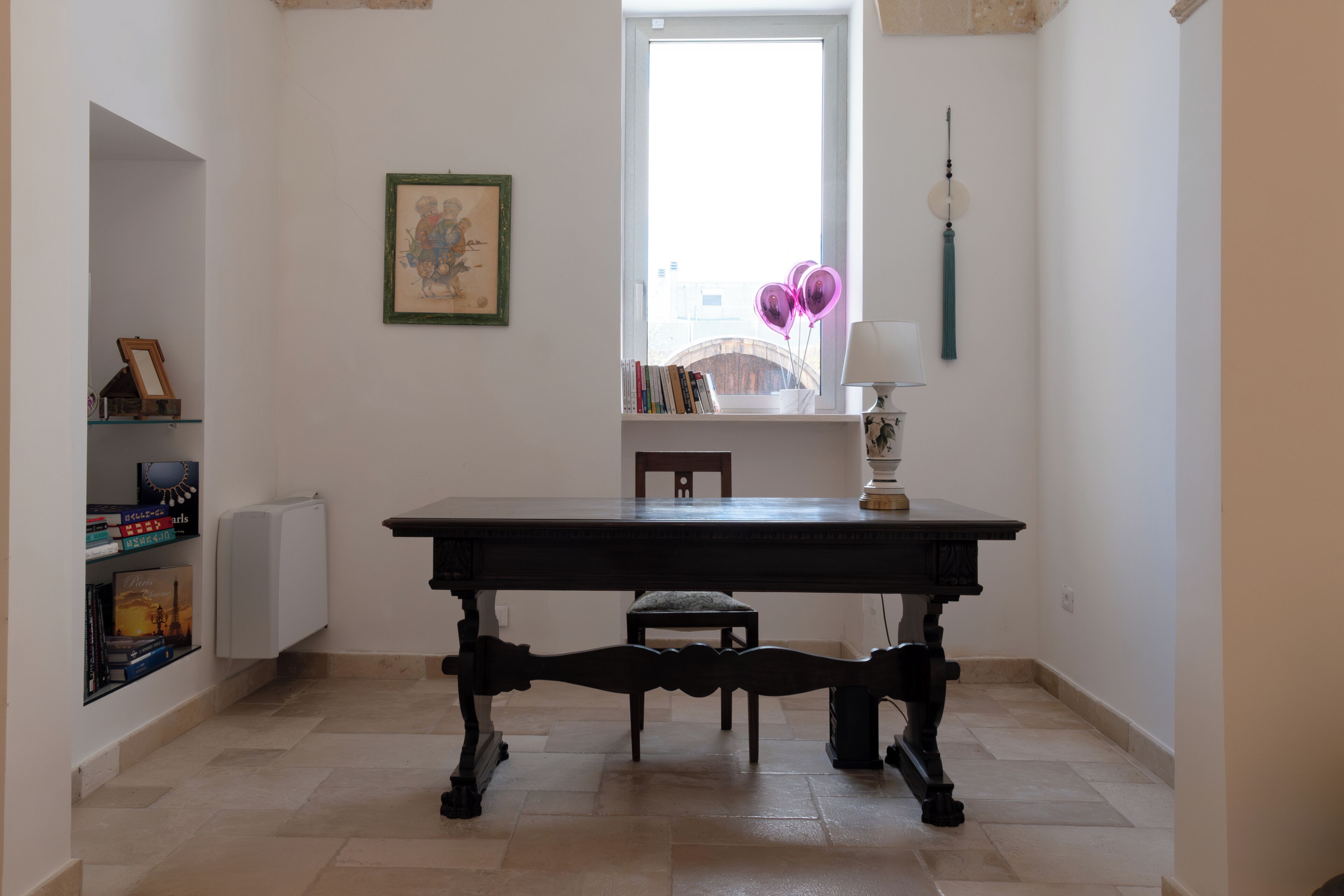 Una habitación con una mesa de madera oscura, una ventana con vistas, una estantería y una lámpara.