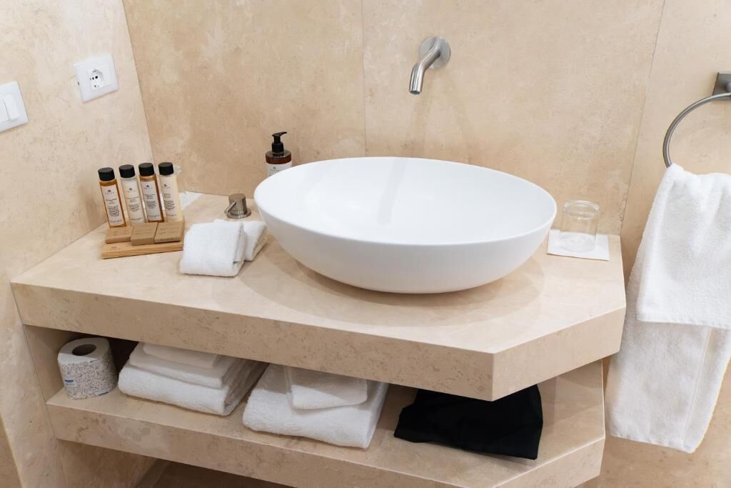 A bathroom with a large oval sink, a towel rack, a soap dispenser, and a shelf with towels and toiletries.