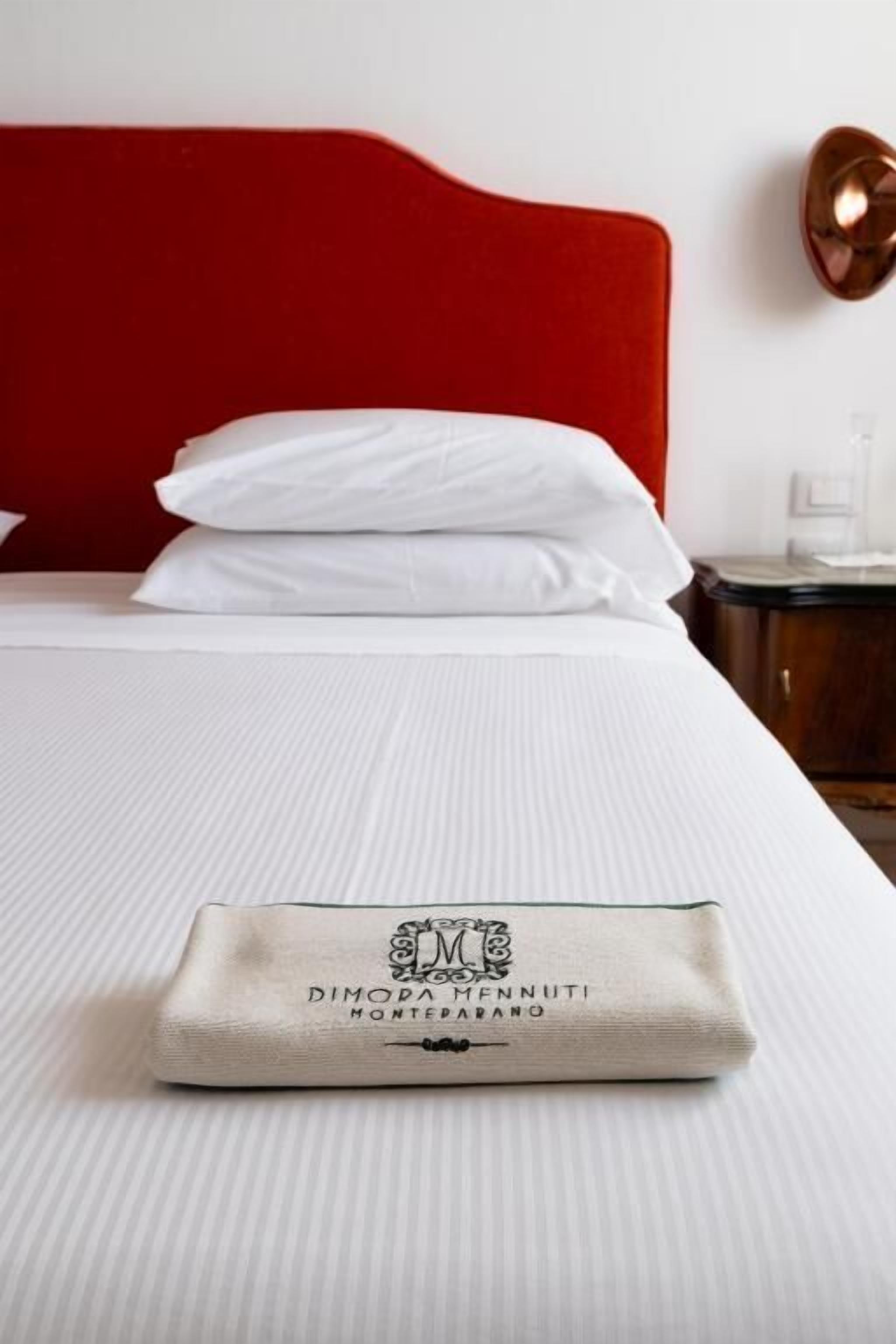 Una cama bien tendida con sábanas blancas, un cabecero rojo y una toalla doblada con un logo.