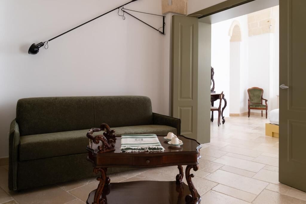 Une pièce avec un canapé vert, une table basse en bois sur laquelle se trouvent un plateau et des tasses, une lampe fixée au mur et une porte qui donne sur une autre pièce.