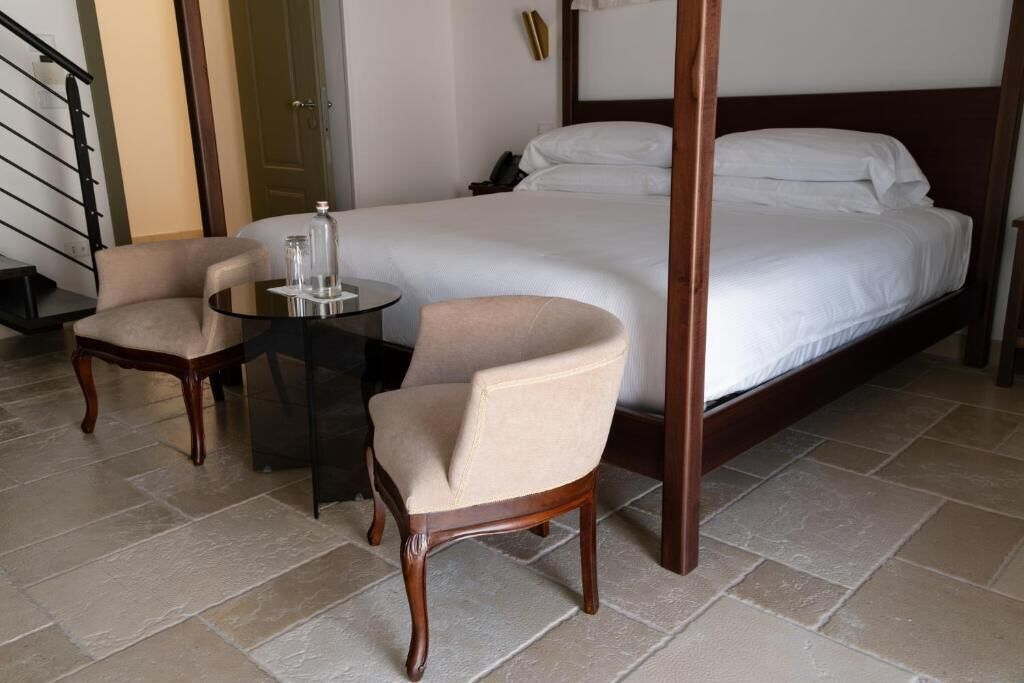 Habitación de hotel con cama con dosel, una pequeña mesa redonda con sillas y una mesita de noche con lámpara.
