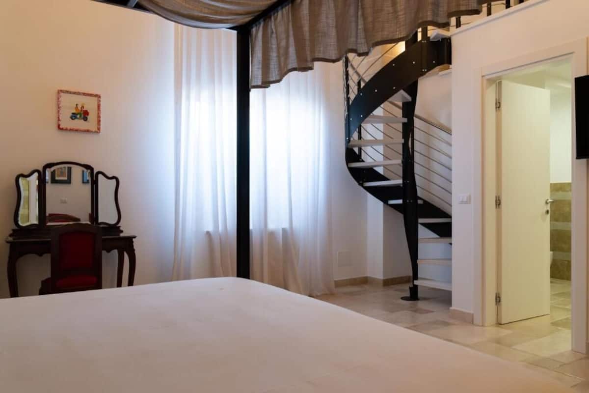 A bedroom with a bed, a desk with a chair, a picture frame, a window with curtains, and a spiral staircase.