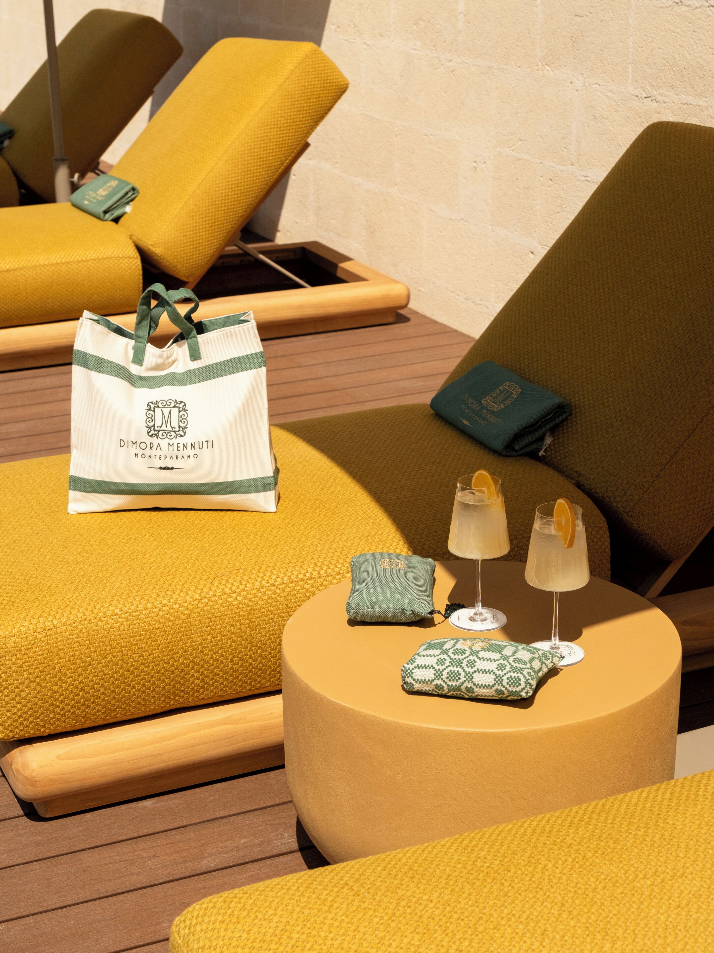 A sunlit outdoor seating area with yellow lounge chairs, a round table with drinks, and a white tote bag.