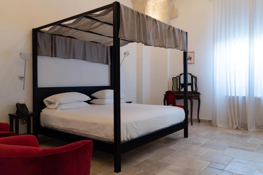 Een hemelbed met baldakijn, een nachtkastje met lamp, een stoel en een tafeltje met een fotolijst.