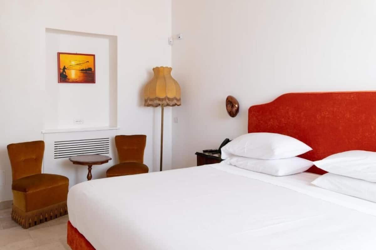 A hotel room with a bed, two chairs, a lamp, a painting, and a red accent wall.