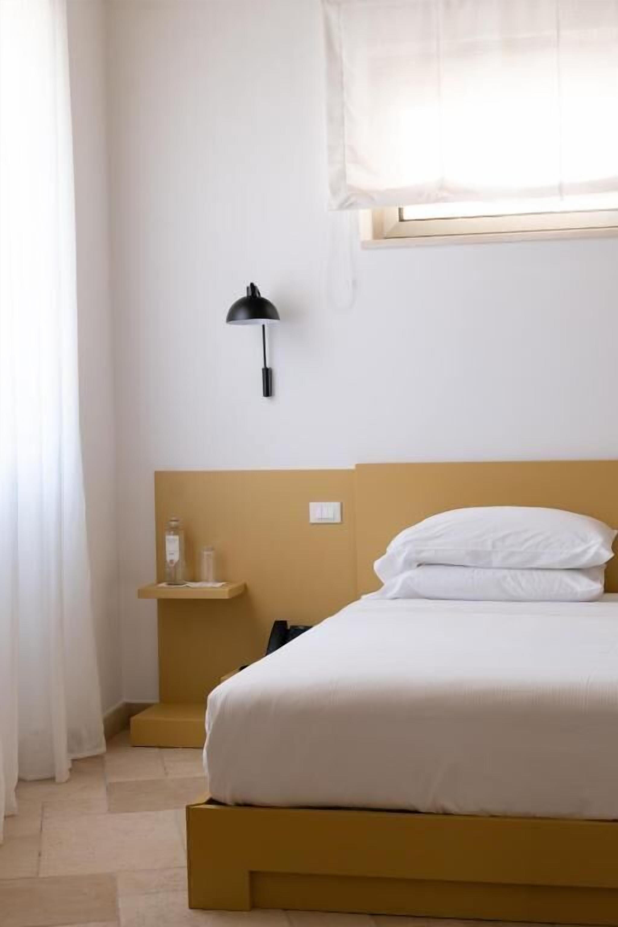 A bed with white bedding, a bedside lamp, and a small shelf with a bottle.