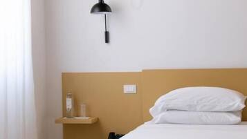 A bed with white bedding, a bedside lamp, and a small shelf with a bottle.