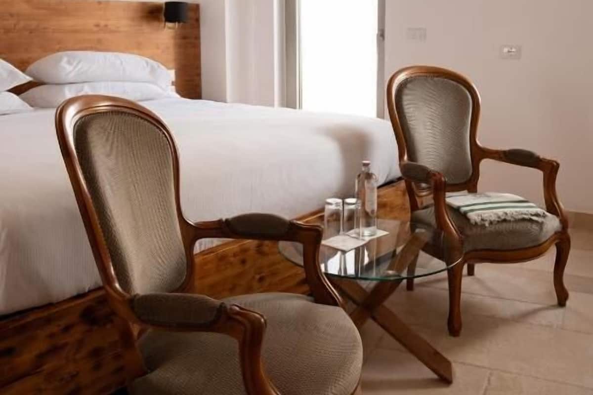 A hotel room with a bed, two wooden chairs, a glass table, and a painting on the wall.