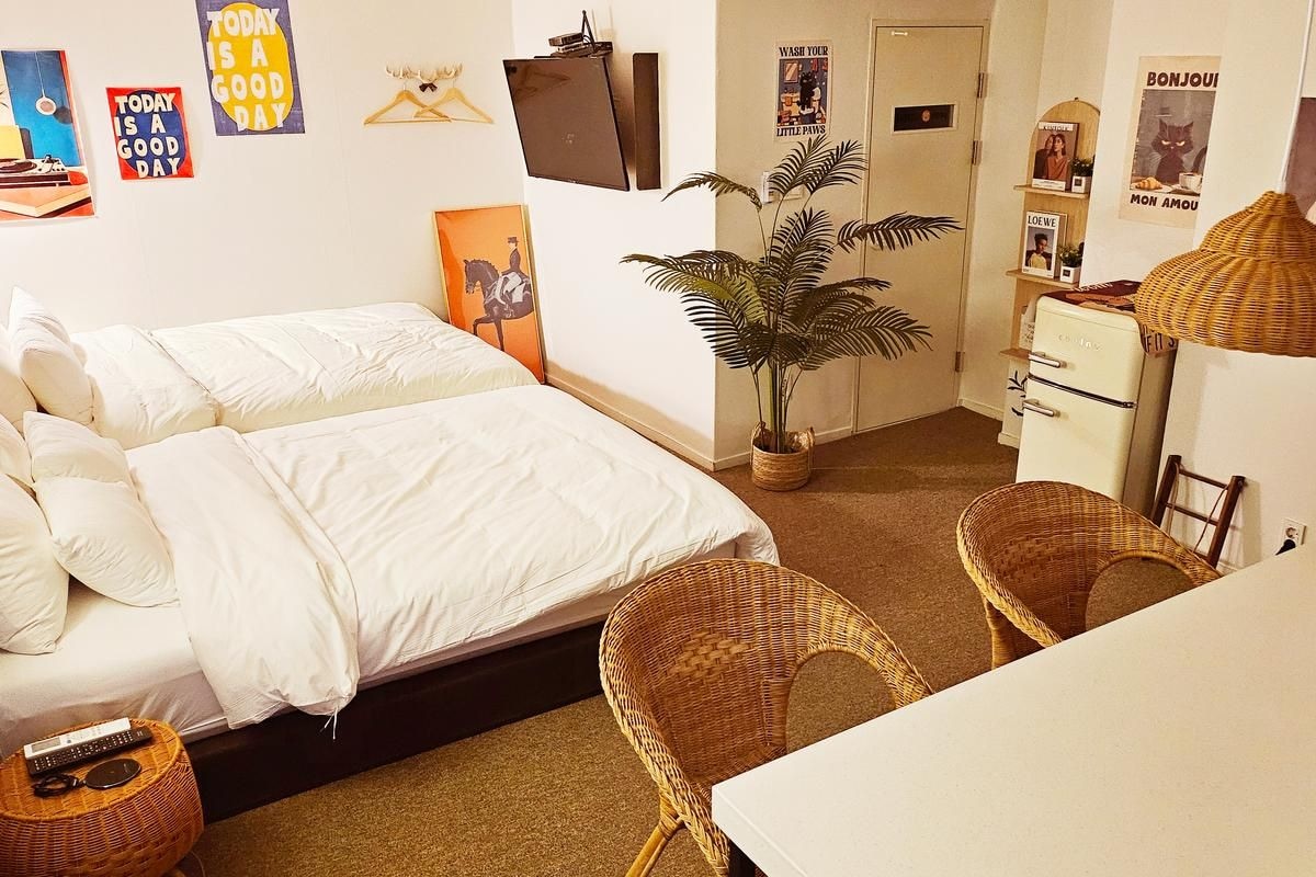Un dormitorio con cama, dos sillas de mimbre, una mesita, una planta en maceta y varias decoraciones en la pared.