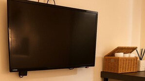 Televisão de tela plana na parede, cesta de vime com velas, mesa pequena e um banquinho tecido sobre um tapete listrado.