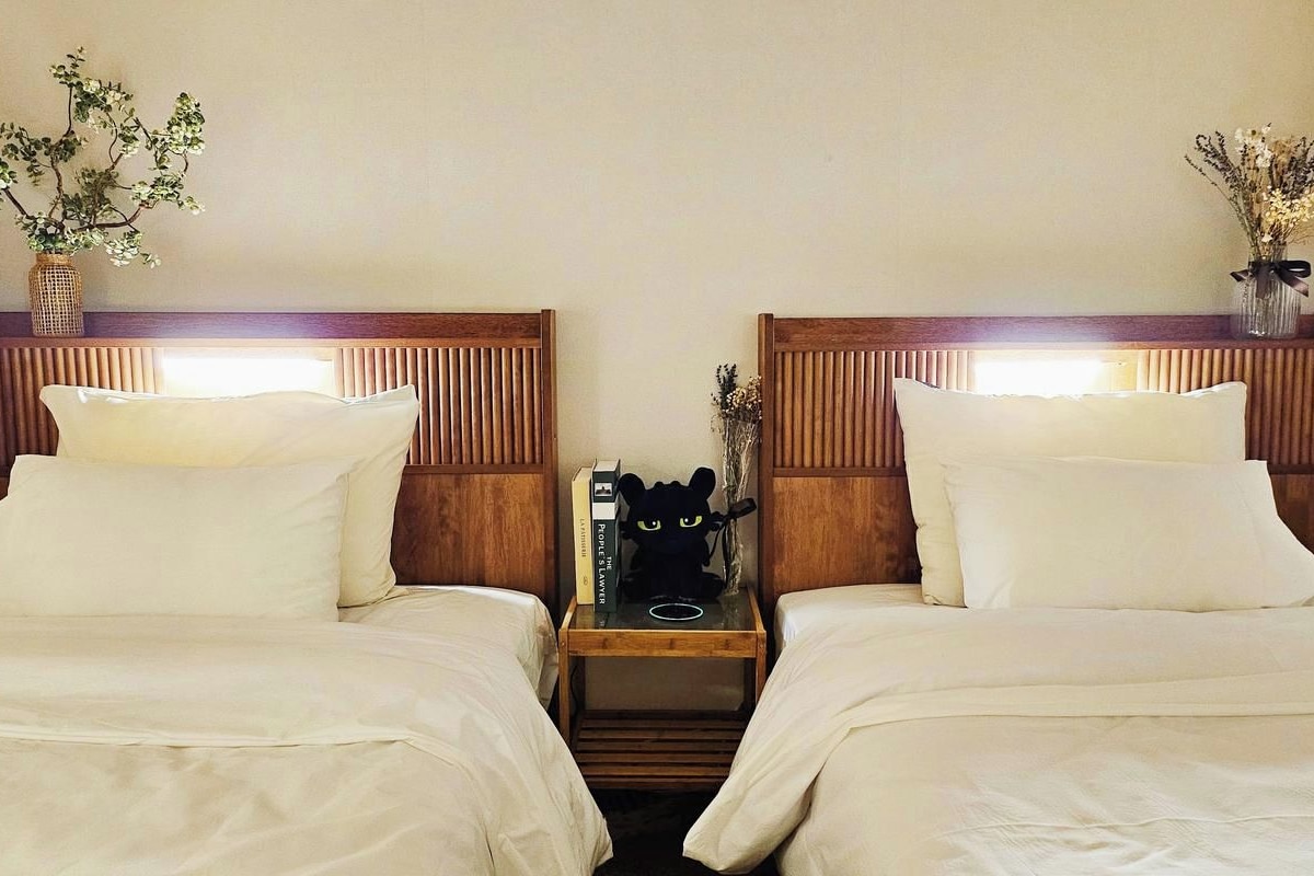 Habitación de hotel con dos camas, cabeceros de madera, ropa de cama blanca, una mesita de noche con lámpara y jarrones decorativos.