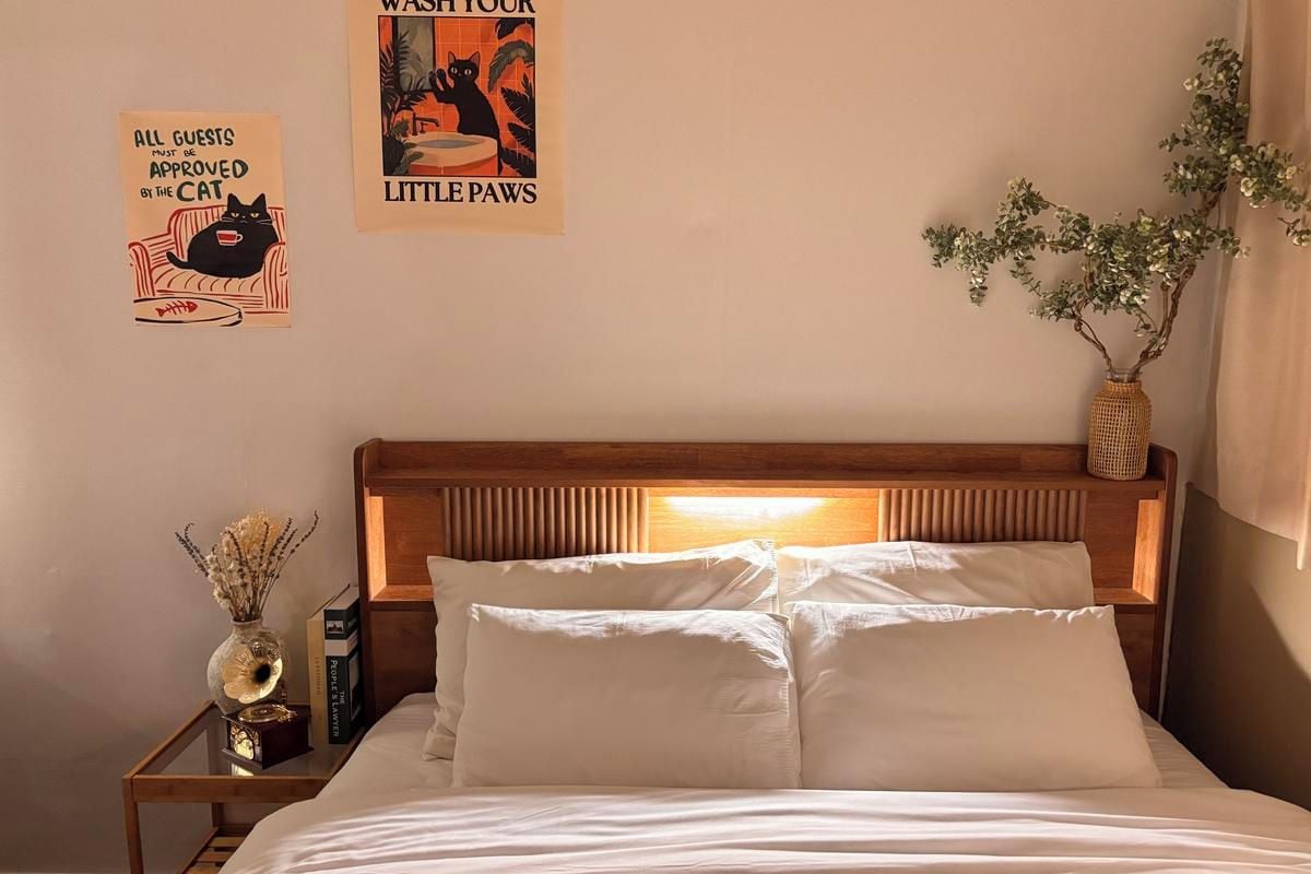 A neatly made bed with white linens, a bedside table with a vase of dried flowers, and two posters on the wall.