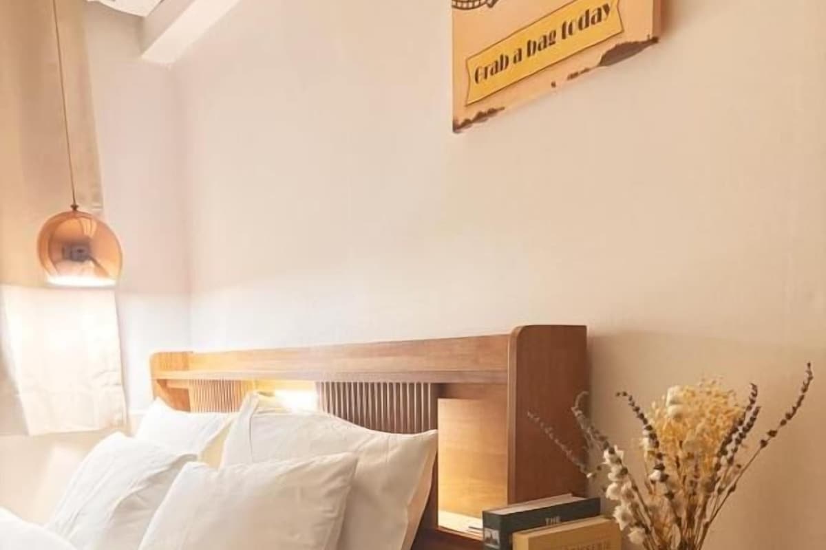 Una cama bien hecha con sábanas blancas, una mesita de noche con un gramófono, un jarrón con flores secas y un letrero en la pared.