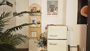 Una habitación con un refrigerador blanco de estilo vintage, un estante de madera con libros y una planta pequeña, un cartel con un gato y una planta verde grande al frente.