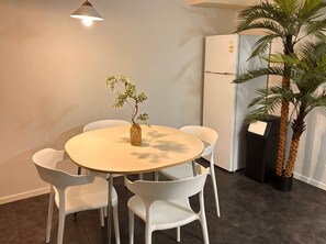 Un comedor con una mesa redonda, sillas blancas, una pequeña planta en maceta, un refrigerador y una planta alta en maceta.