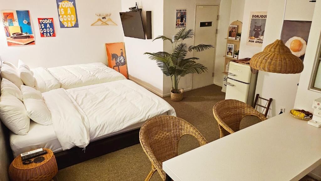 A bedroom with a bed, two chairs, a desk, a plant, and a refrigerator.