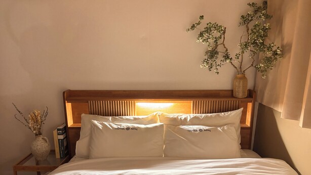 Cama bem arrumada com roupa de cama branca, cabeceira de madeira, mesa de cabeceira com um vaso de flores secas e uma planta fixada na parede.