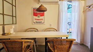 Uma área de refeições com uma mesa, quatro cadeiras de vime, um candeeiro fixado na parede e um cartaz de um produto enlatado.