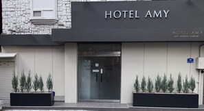 L’entrée d’un hôtel, avec une enseigne indiquant « HÔTEL AMY » et un panneau bleu indiquant le numéro de l’immeuble, soit le 10.