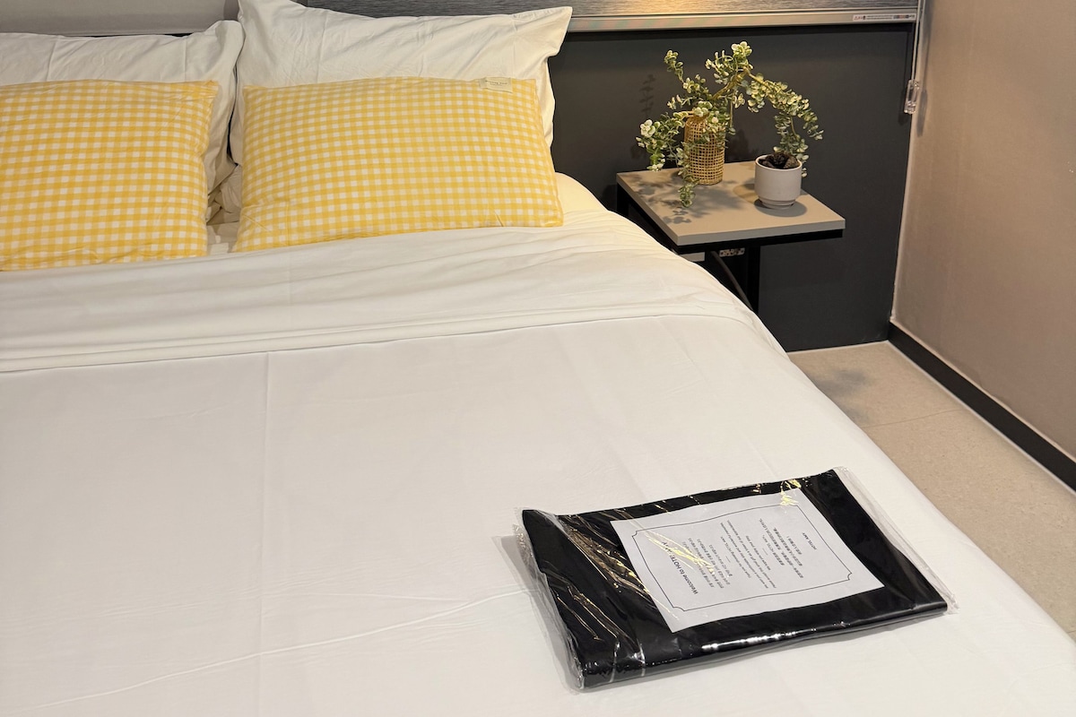 A neatly made bed with white and yellow checkered pillows, a bedside table with a plant, and a wall-mounted light fixture.