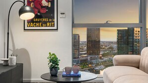Un salon moderne avec vue sur la ville, une affiche encadrée, une table avec des livres et une plante, et une lampe.