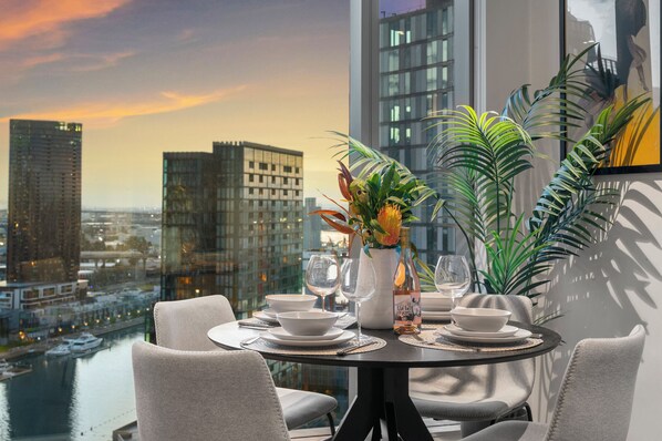 Una mesa de comedor con un florero, copas de vino y platos para dos, con vistas a un paisaje urbano con edificios y una masa de agua.