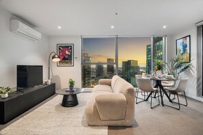 Ein modernes Wohnzimmer mit einer Couch, einem Esstisch und Stühlen, einem Flachbildfernseher und Blick auf die Stadt durch große Fenster.