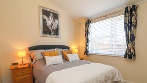 A neatly made bed with a headboard, a bedside table with a lamp, a framed picture on the wall, and a window with curtains. - Banksy's Retreat (Ilfracombe)