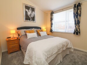A neatly made bed with a headboard, a bedside table with a lamp, a framed picture on the wall, and a window with curtains. - Banksy's Retreat (Ilfracombe)