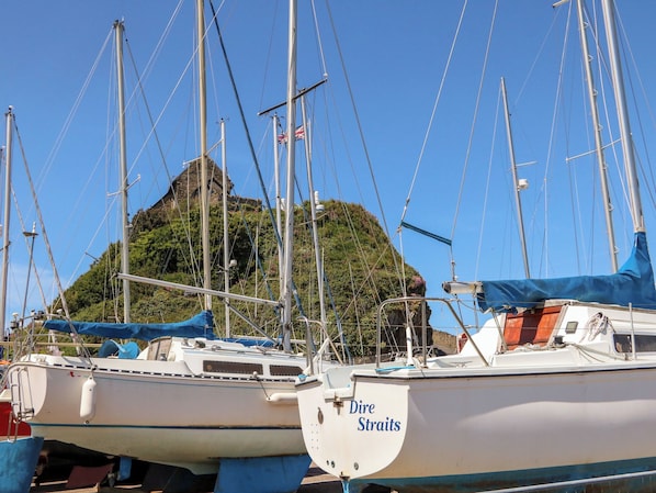 A marina with several sailboats docked, a stone building in the background, and a hill with greenery.