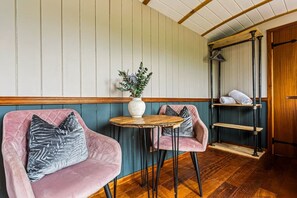 Una habitación con dos sillones tapizados en rosa, una mesita redonda pequeña, un jarrón blanco con flores y una estantería de madera con toallas.