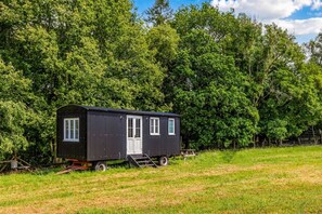Ein kleiner, dunkel gefärbter Wohnwagen mit einer Haustür, Fenstern und einer Rampe, gelegen in einem grünen Feld mit dichtem Baumbestand im Hintergrund.