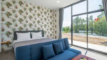 A bedroom with a bed, a blue sofa, a wooden coffee table with flowers, and a large window with a view of trees and a road.