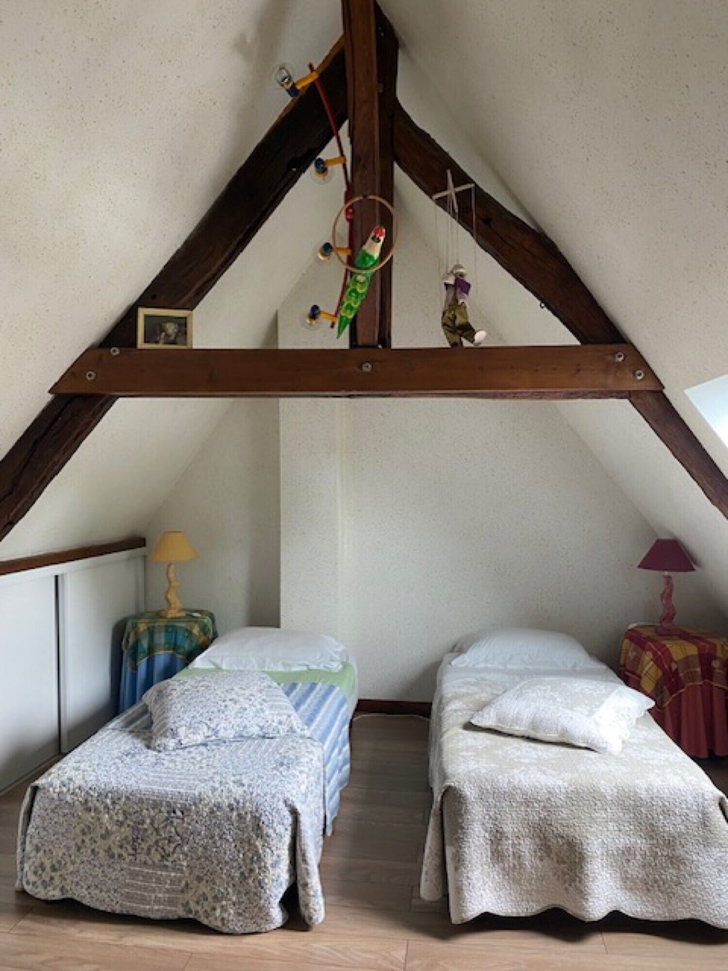 Een knus zolderkamertje met twee bedden, houten balken, een hanglamp en een klein raam.