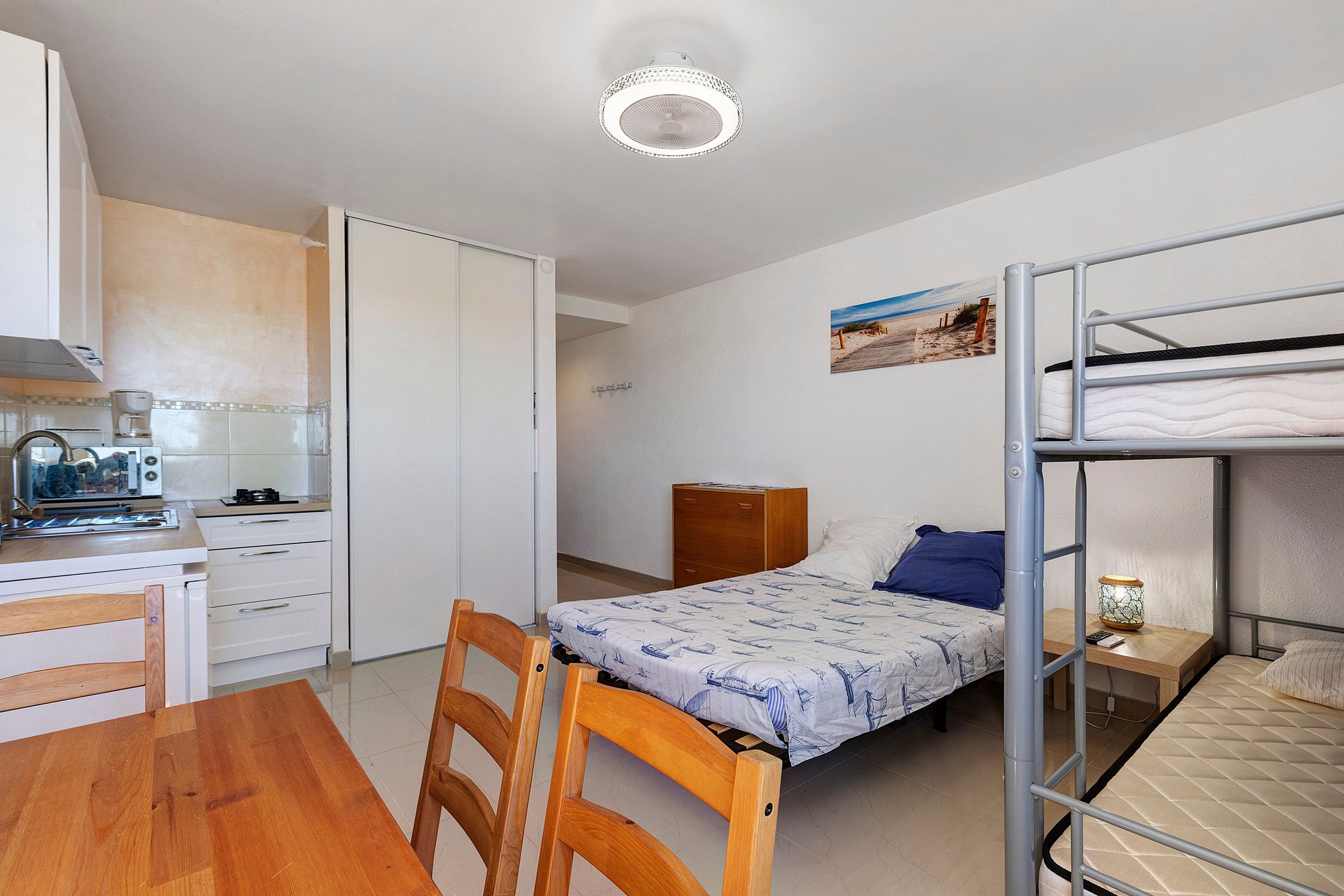 Een kleine kamer met een stapelbed, een bureau, een stoel, een kleine kitchenette en een bed met een bedspread met dessin.