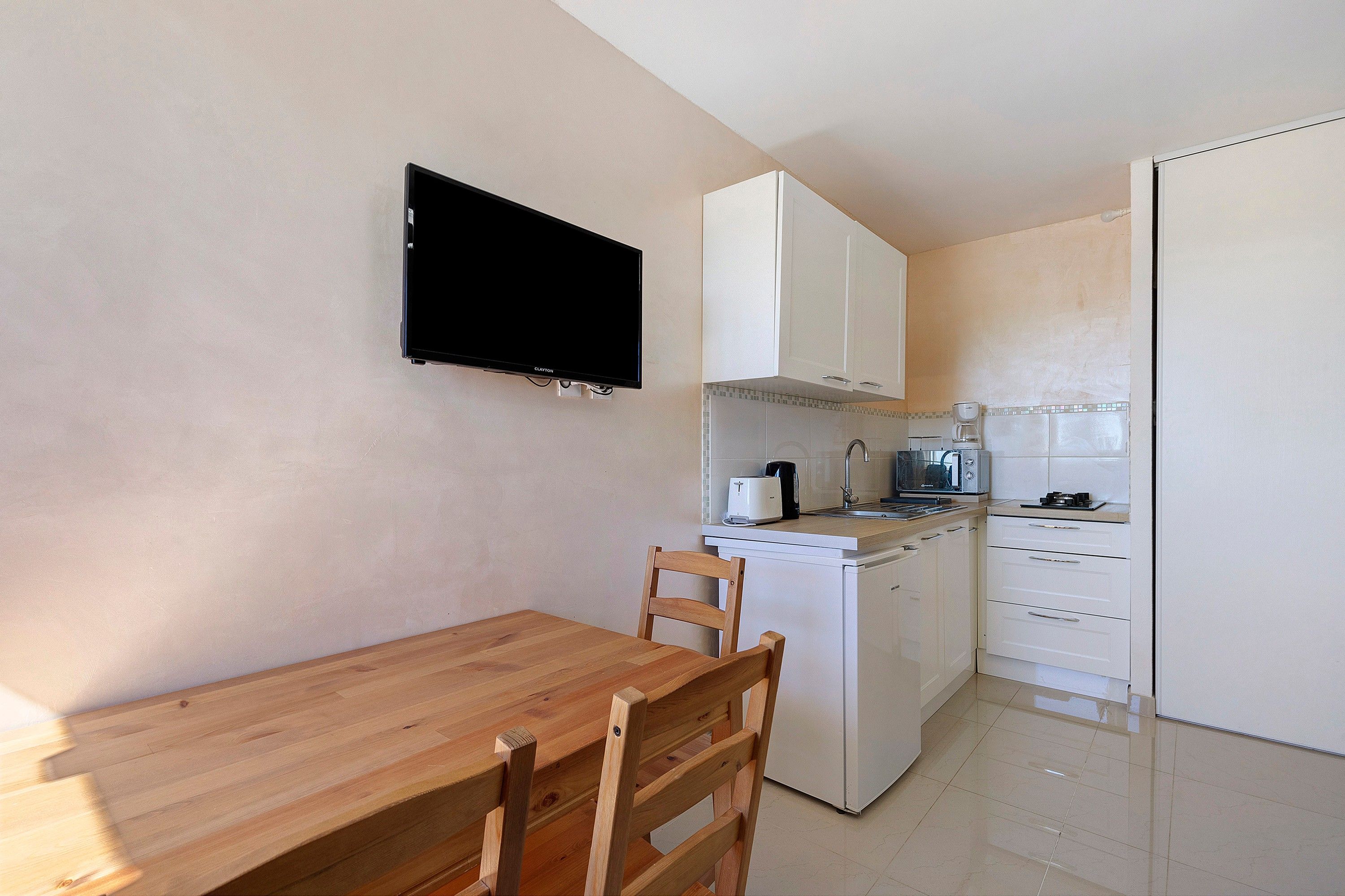 Een kleine keuken met een houten tafel en stoelen, een flatscreen televisie bevestigd boven het aanrecht en een magnetron.
