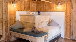 Interior de una cabaña de madera con un banco de piedra, almohadones, un plaid de piel y una cafetera.