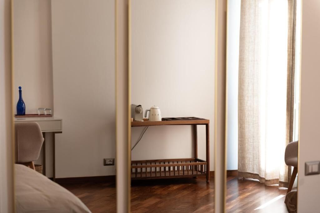 Una habitación con cama, escritorio, una mesita con tetera y ventana con cortinas.