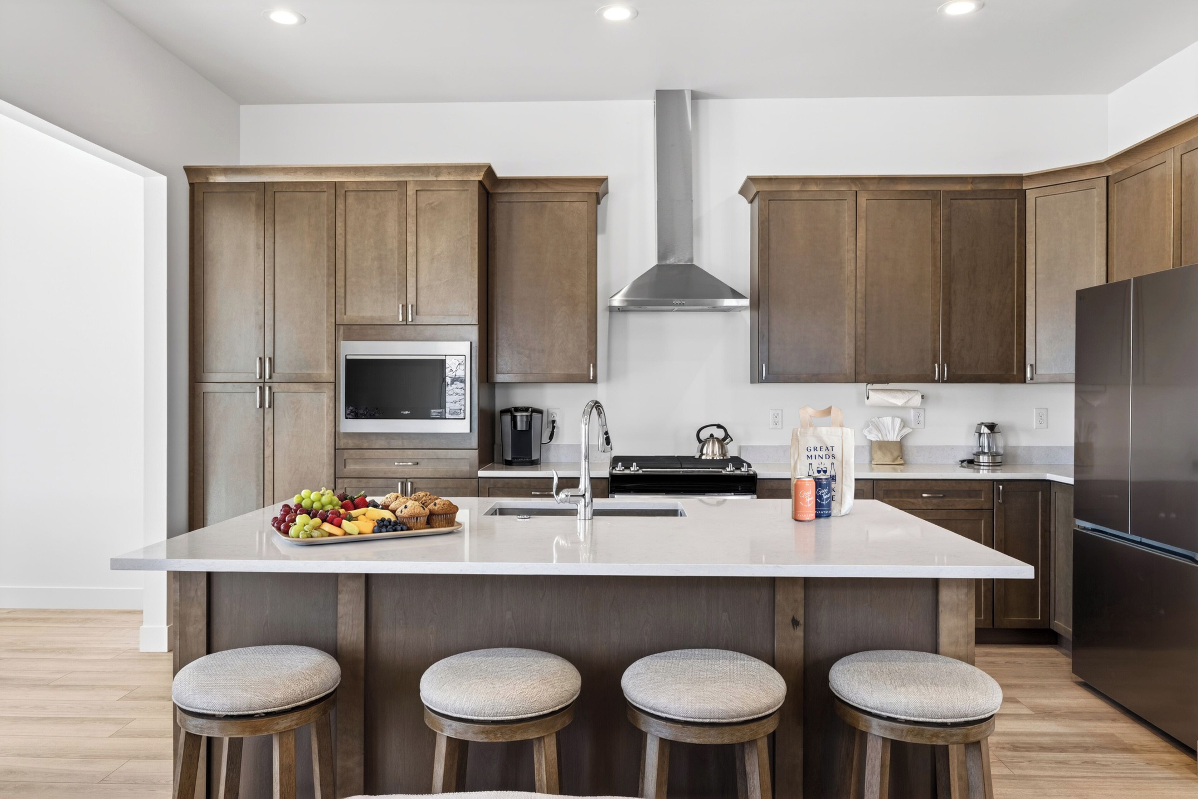 A modern kitchen with wooden cabinets, a stainless steel range hood, a central island with stools, and a microwave.