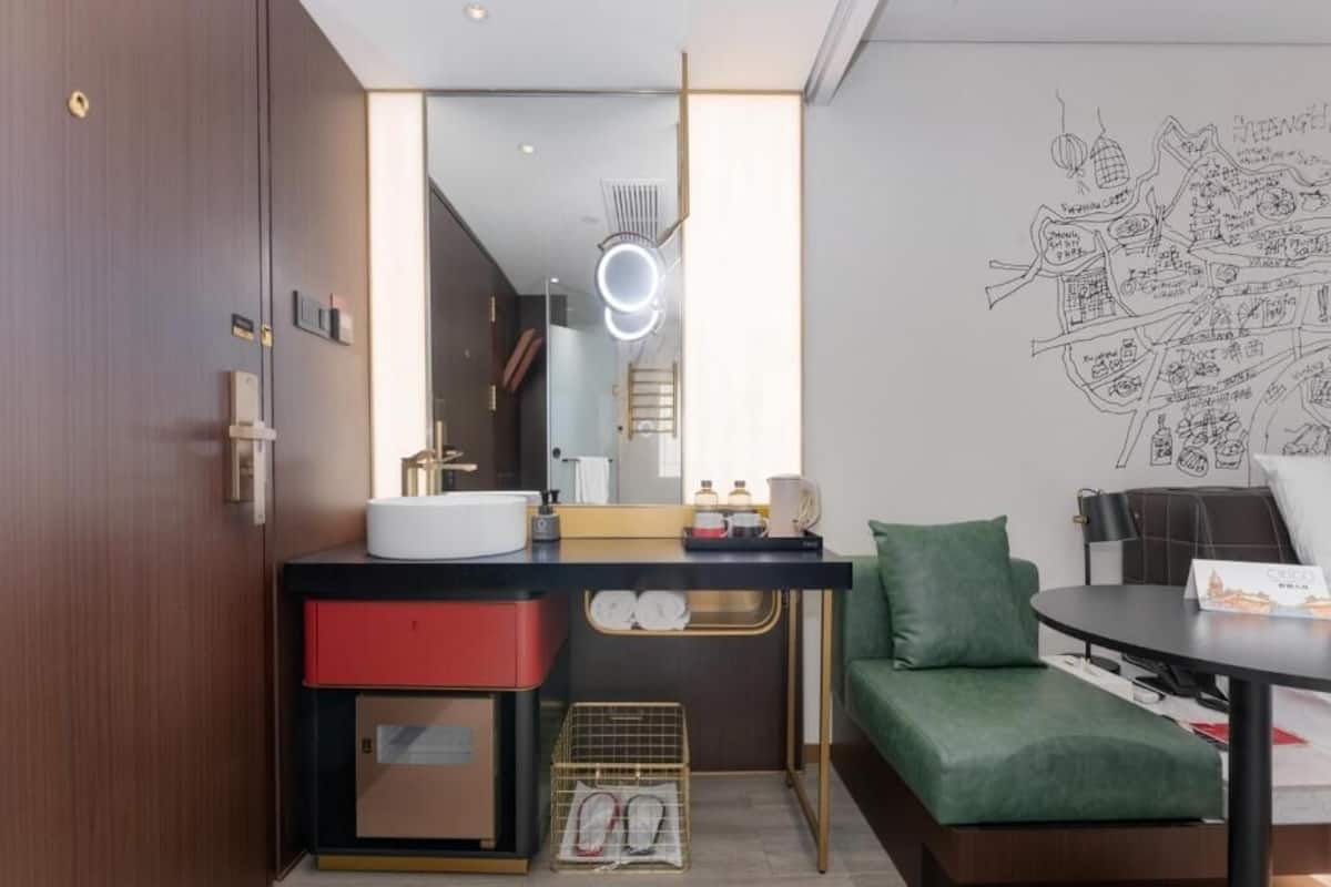A modern hotel room with a large mirror, a sink, a green sofa, and a wall with a cityscape sketch.
