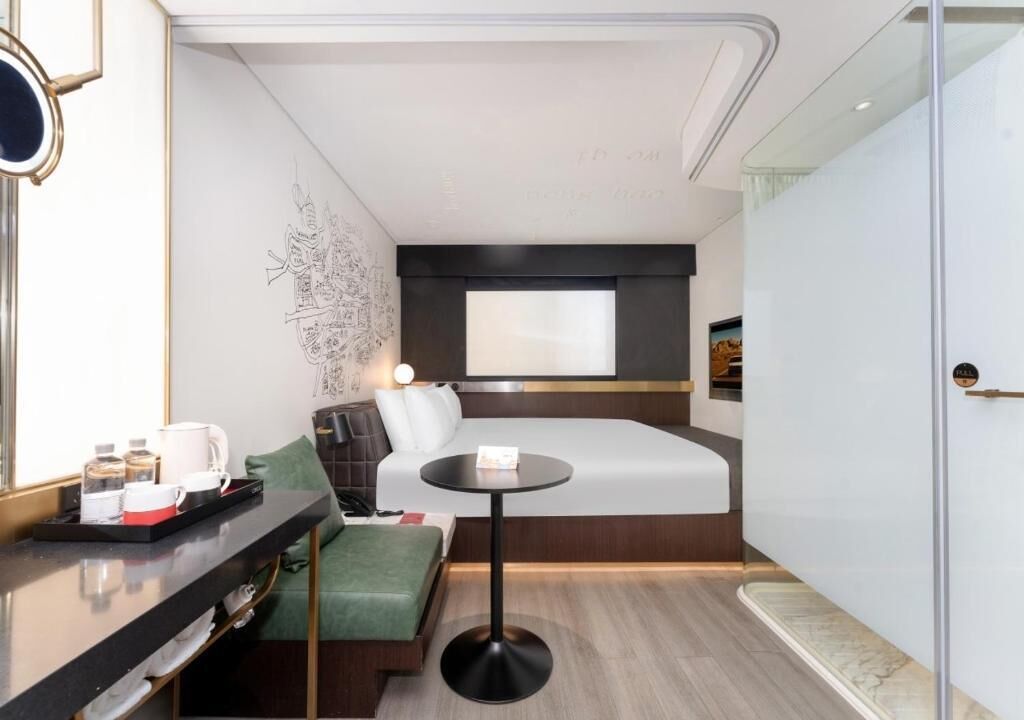 Una camera d'albergo compatta con un letto, un tavolino rotondo, una poltrona e una scrivania con un vassoio di oggetti, oltre a una parete decorata con un motivo ornamentale.