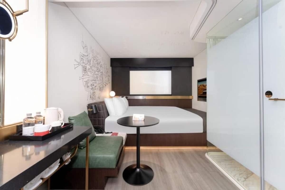 Ett kompakt hotellrum med en säng, ett litet runt bord, en soffa, ett skrivbord med ett bricka fullt med diverse föremål och en vägg med ett dekorativt mönster.