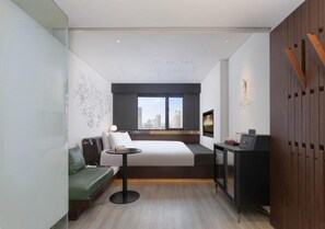 A modern hotel room with a bed, a small desk, a seating area, and a city view.