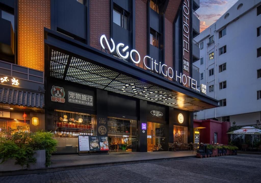 Un hotel con fachada moderna, un llamativo letrero de neón y un restaurante visible con terraza.
