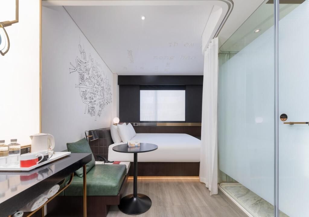 Una camera d'albergo moderna con un letto, un tavolino, una panca verde e un divisorio in vetro.