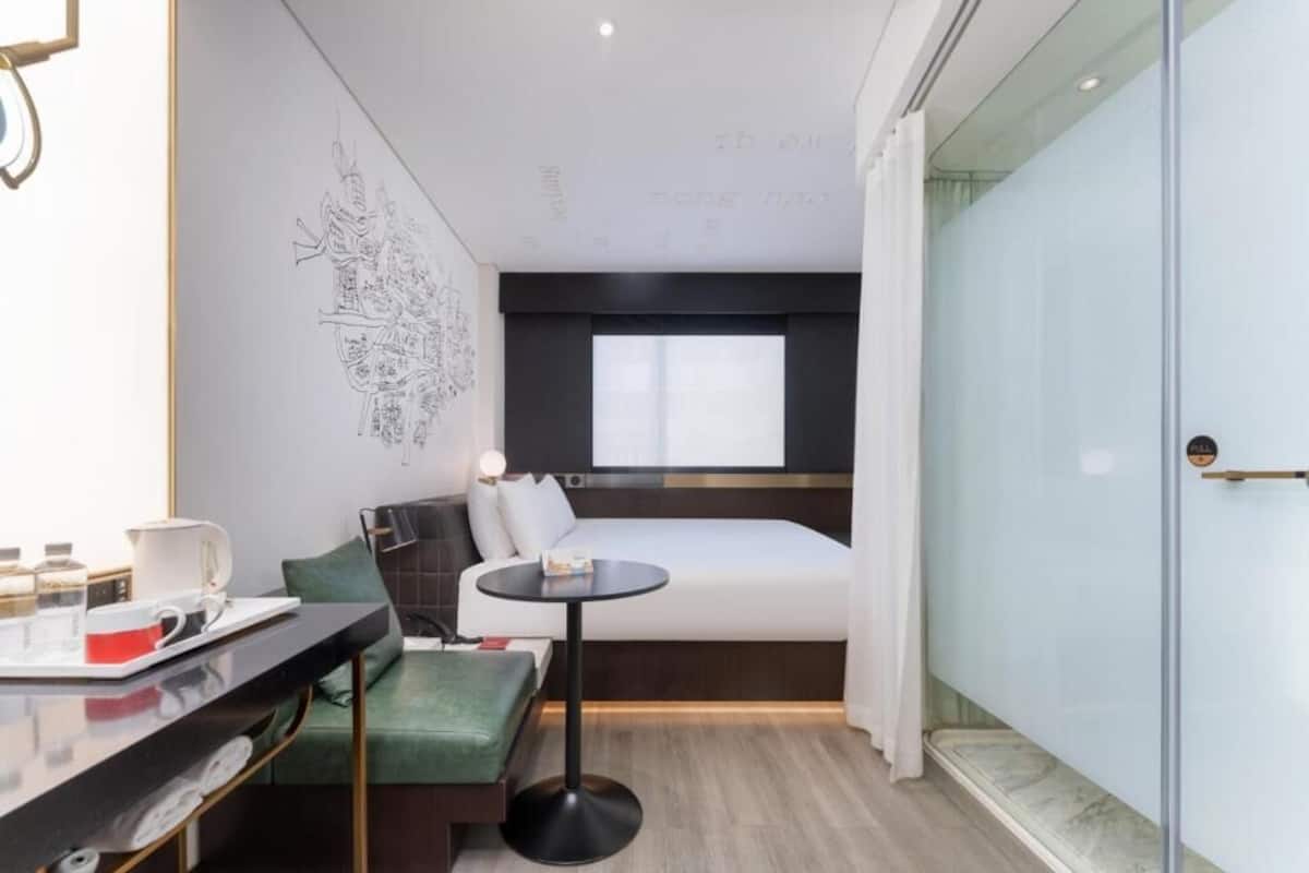 Ett modernt hotellrum med en säng, ett litet bord, en grön bänk och en glasvägg.