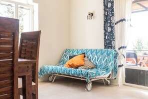 Ein Zimmer mit einem Holzstuhl, einem gemusterten Sofa, einer kleinen Wandleuchte und einem Fenster mit Vorhängen.
